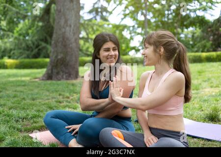 Zwei lächelnde junge Mädchen in Sportswear, die sich gegenseitig hochkitschen. Sie sitzen im Park auf dem Gras. Platz für Text. Stockfoto