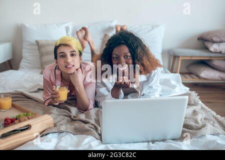 Weibliches Paar liegt auf dem Bett mit Laptop und Frühstück Stockfoto