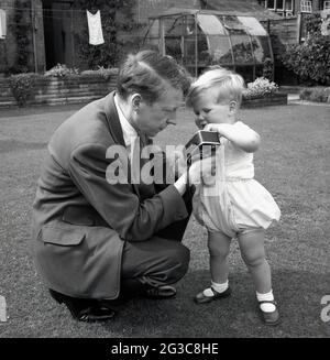 In den 1950er Jahren, historisch, draußen in einem Garten, kniete ein Vater in Anzug und Krawatte nieder und zeigte seinen kleinen Sohn, seine kleine, handgehaltene 8-mm-Cine-Kamera, England, Großbritannien. Solche leichten Cine-Kameras mit 8mm-Film waren in dieser Ära ein Popula-Hobbyr, der verwendet wurde, um sogenannte „Heimfilme“ zu drehen, die Aufzeichnungen über das gewöhnliche Leben der Menschen drehten. Stockfoto