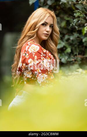 latina mit blondem Haar, die an einem sonnigen Tag neben einer Gartenpflanze steht und lässige Bluse, Lifestyle und Natur trägt Stockfoto