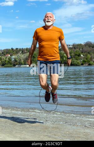Aktiver älterer Mann, der am Ufer des Flusses am Strand trainiert und am Seil springt. Stockfoto
