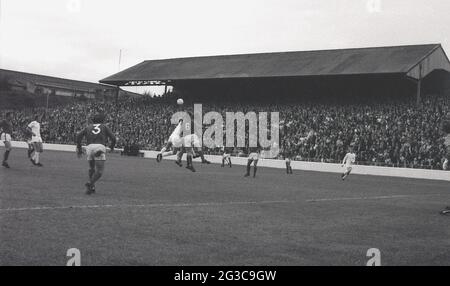 1970s, historisch, ein Fußballspiel, Millwall verus Charlton, auf ihrem Boden, The Den, Cold Blow Lane, Surrey Docks, East London, England, Großbritannien, wo der FC Millwall von 1910 bis 1993 spielte. Das Bild zeigt die Spieler, die um den Ball kämpfen, und die halbwegs überdachte Terrasse mit Teiltribünenabdeckung, die Nordterrasse, die voller Zuschauer ist. Das von Archibald Leitch entworfene „The Den“ war 83 Jahre lang die Heimat des FC Millwall und ein feindseliger, einschüchternder Austragungsort gegnerischer Teams. Durch die Luftangriffe von WW2 schwer beschädigt, wurde es mit Hilfe von Unterstützern wieder aufgebaut und 1944 wiedereröffnet. Stockfoto