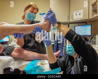 Multirassische Krankenschwestern, die Gesichtsmasken tragen, untersuchen eine Verletzung an der Ferse einer weiblichen Patientin in einer Krankenschwesterklinik in Aliso Viejo, CA. Stockfoto