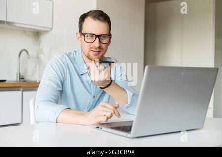 Ein intelligenter junger Mann sitzt am Küchentisch und arbeitet fernab von zu Hause aus, nutzt einen Laptop und nimmt Sprachnachrichten auf, ein männlicher Unternehmer nutzt eine Spracherkennungs-App auf seinem Mobiltelefon Stockfoto