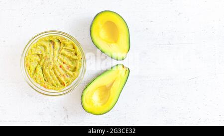 Zwei Avocado-Hälften und eine Glasschale mit vorbereiteter Guacamole auf einem Schreibtisch mit weißem Stein, Blick von oben für Text rechts Stockfoto