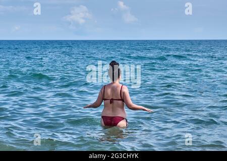 Eine junge asiatische Frau mit Frisur von Dreadlocks auf schwarzem Haar, in einem roten Badeanzug, zu Fuß ins Meer. Blick von hinten. Sommerlandschaft. Stockfoto