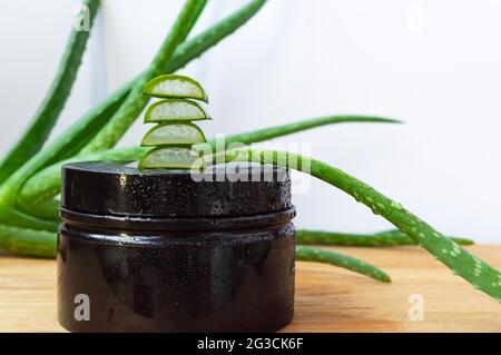 Ganze und in Scheiben geschnittene Aloe Vera Blätter und kosmetisches schwarzes Glas mit Wassertropfen auf einem hölzernen Hintergrund. Naturkosmetik Stockfoto