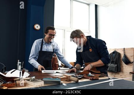 Zwei junge Lederhandwerker, Freude und Witz während der Arbeit in der Werkstatt, positive Lederschneider, haben Spaß an der Arbeit. Kleines Familienunternehmen Stockfoto