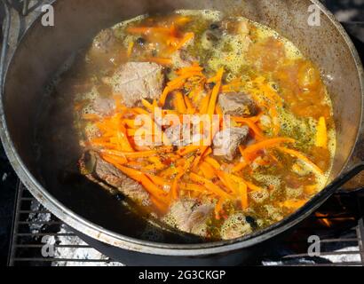 Kochen traditioneller orientalischer Pilaf in einem Kessel über einem Feuer. Geschmorte Fleischstücke und Zwiebeln, Karotten und Gewürze werden mit kochendem Wasser und s gegossen Stockfoto