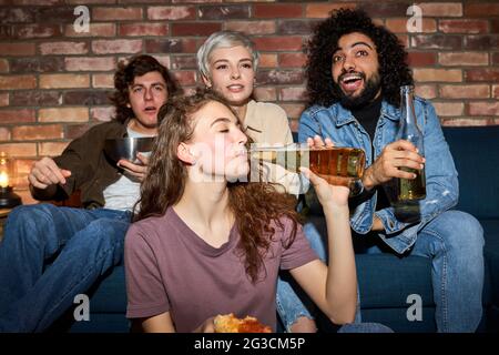 Junge Freunde schauen fernsehen, Comedy-Show oder Film, essen Snacks und trinken Getränke, sitzen auf der gemütlichen Couch zu Hause, diverse Jungs und Damen genießen Stockfoto