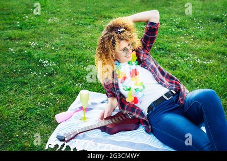 Glücklich lachend lockiges Haar Frau entspannen nach Ukulele Gitarre spielen auf üppigem grünen Gras in einem Stadtpark bei einem Picknick. Sommer positive Stimmung. Spaß haben Stockfoto