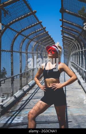 Eine blonde weibliche Model, im Pony-Schwanz trägt einen transparenten roten Visierhut, Sport-BH und Shorts, die mit ihren beiden Händen auf ihrer Hüfte posieren. Stockfoto