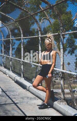 Eine blonde weibliche Model, in einem Fitness-Outfit, das sich gegen einen Metallzaun lehnt und eine Hantel hebt. Sie trägt einen schwarzen Sportbra und schwarze Shorts. Stockfoto