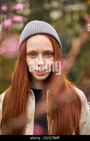 Porträt einer wunderschönen kaukasischen Frau, die am Frühlings- oder Herbsttag im Freien posiert, zwischen blühenden Bäumen steht und die Kamera anschaut, rothaarige Weibchen ist posinisch Stockfoto