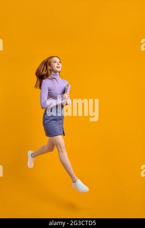 Optimistische Rotschopf-Dame beim Springen am Wochenende, halten Notebooks auf dem Weg nach Hause in der Luft in den Händen, tragen lässiges Hemd und Rock isoliert gelb orange Rückenrock Stockfoto