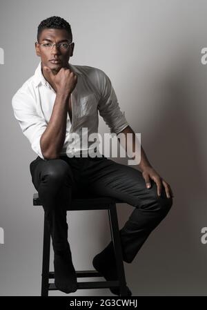 Ein junger schwarzer Mann, der eine Brille trägt, ein Modelmodell, der auf einem Holzhocker sitzt und mit der Hand bis zum Kinn posiert, ein ernstes Porträt. Stockfoto