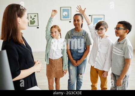 Vielfältige Gruppe von Kindern, die die Hände heben, während sie einer Expertin in der Galerie für moderne Kunst zuhören Stockfoto