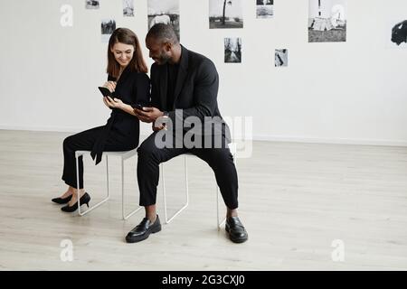 Minimalistisches Hochwinkelporträt eines eleganten, schwarz getragenen Paares mit gemischter Rasse, das Smartphones in einer modernen Kunstgalerie verwendet, um Platz zu kopieren Stockfoto