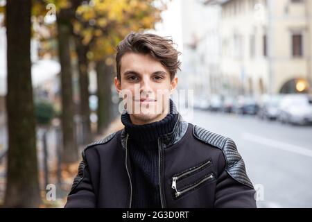 Porträt eines attraktiven jungen kaukasischen Mannes, der an einem bewölkten Herbsttag in der Stadt ist, ist ein hübscher Kerl mit braunen Haaren. Gelassen, lächelnd und Stockfoto