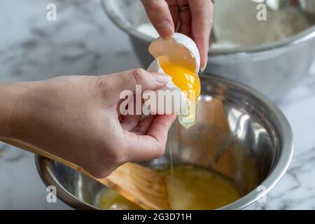 Die Hand einer Frau, die mit dem gelben Eigelb ein Ei aufbricht, über einer runden Metallschüssel mit natürlichem Licht und Marmoroberfläche Stockfoto