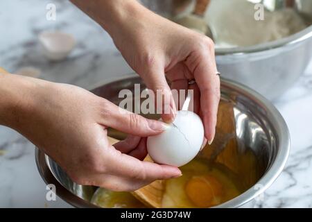 Die Hand einer Frau, die ein Ei über einer gewickelten Metallmischschüssel mit natürlichem Licht aufbricht Stockfoto