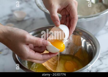 Die Hand einer Frau, die mit dem gelben Eigelb ein Ei aufbricht, über einer runden Metallschüssel mit natürlichem Licht Stockfoto