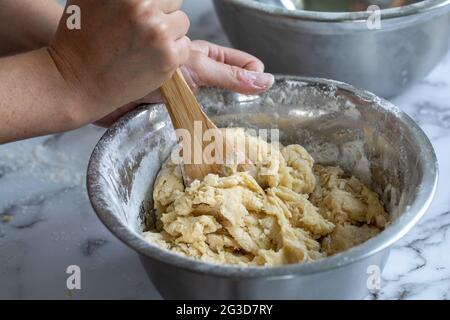 Nahaufnahme der Hände einer Frau, die einen Holzlöffel hält, der Zutaten in einer runden Metallschale auf einer weißen Marmoroberfläche mischt Stockfoto