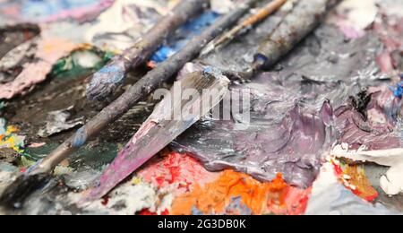 Die Palette eines Künstlers oder Malers ist mit einer gemischten, dicken Ölfarbe bedeckt. Palettenmesser und Pinsel liegen diagonal über dem meist violetten, blauen A Stockfoto