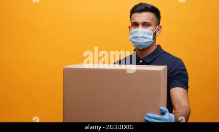 Lieferung Mann trägt Maske und Handschuhe mit einem Karton mit gelbem Hintergrund. Paketzustellung während des Pandemiekonzepts. Stockfoto