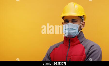 Porträt eines jungen indischen Ingenieurs mit einem gelben Schutzhelm und einer Maske, die im Studio mit gekreuzten Armen auf gelbem Hintergrund steht. Isolierter Mann. Nahaufnahme. Stockfoto