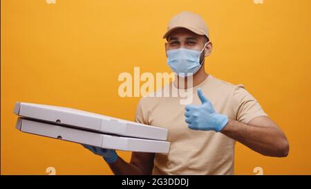 Der Lieferjunge trägt eine Maske und Handschuhe, hält Pizzakisten und macht die Daumen hoch und lächelt. Konzept der Heimlieferdienste während der Covid-19. Isolierter Mann mit gelbem Hintergrund Studio. Stockfoto