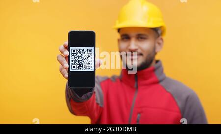 Ein Mann mit gelbem Helm und Uniform zeigt der Kamera das Handy mit QR-Code auf dem Bildschirm. Studioaufnahme mit gelbem Hintergrund. Digitales Bezahlkonzept. Stockfoto