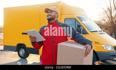Der Lieferjunge trägt eine Uniform und eine Kappe mit einem Pappkarton, der auf einem iPad nach Adresse sucht und hinter ihm einen gelben Van hat. Konzept des Paketzustelldienstes. Stockfoto