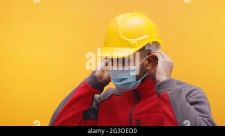 Porträt eines jungen indischen Ingenieurs, der einen gelben Schutzhelm trägt und eine im Studio stehende Maske mit gelbem Hintergrund aufsetzt. Konzept der Pandemie. Isolierter Mann. Nahaufnahme. Stockfoto