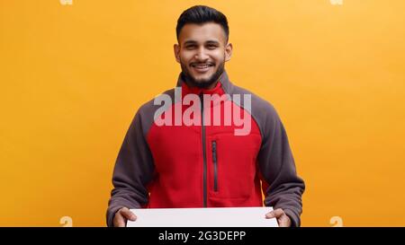 Der Lieferer hält eine Pizzakiste in der Hand. In roter Uniform lächelnd gekleidet. Konzept des Home Delivery Service. Studioaufnahme mit gelbem Hintergrund. Isolierter Mann. Stockfoto