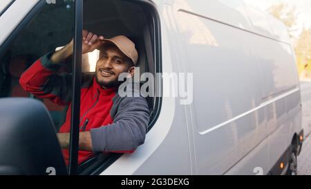 Zustellguy in roter Uniform sitzt im weißen Van und kippt seine Mütze. Fröhlicher Inder, der auf dem Vordersitz sitzt und die Kamera anlächelt. Kurierkonzept. Stockfoto