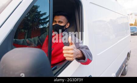 Der Lieferer trägt eine Maske und eine rote Uniform und fährt einen weißen Van, der ein Daumen nach oben trägt. Fröhlicher Inder, der auf dem Vordersitz sitzt und die Kamera anlächelt. Kurierkonzept. Stockfoto