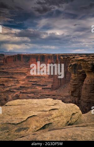 Das Abendlicht leuchtet auf den hoch aufragenden Sandsteintürmen des Monument Basin, während sich Sturmwolken über dieser abgelegenen White Rim Road im Canyonland von Utah sammeln Stockfoto