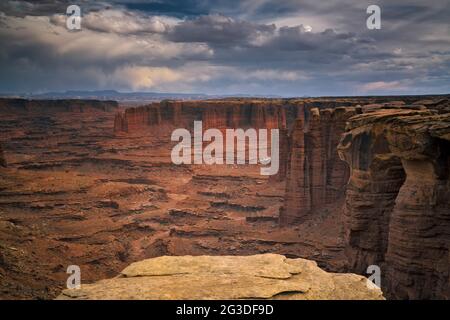 Das Abendlicht leuchtet auf den hoch aufragenden Sandsteintürmen des Monument Basin, während sich Sturmwolken über dieser abgelegenen White Rim Road im Canyonland von Utah sammeln Stockfoto