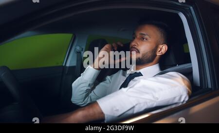 Junger indischer Geschäftsmann sitzt im Auto und telefoniert. In weißem Hemd und Krawatte gekleidet. Die Aufnahme wurde auf dem Parkplatz gemacht. Konzept des Bürogebäudes Parkplatz. Stockfoto