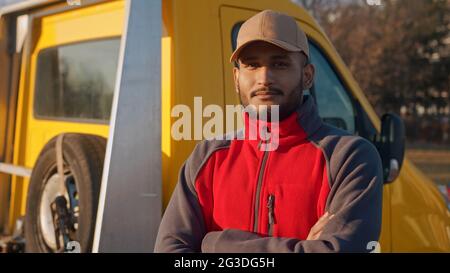 Junger Arbeiter, der mit gekreuzten Armen steht und lächelt. In roter Uniform und einer Mütze gekleidet. Hinter ihm ist ein gelber LKW mit Reserverad zu sehen. Konzept der Automechanik. Nahaufnahme. Stockfoto