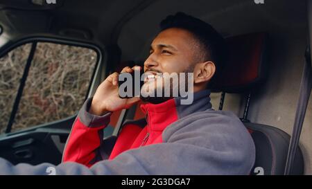 Zustelljunge, der einen Van fährt und telefoniert. Hält ein Telefon in der Hand. In roter Uniform gekleidet. Lächelnd und im Gespräch mit einem Gesprächspartner. Junger Inder. Paketzustellungskonzept. Stockfoto