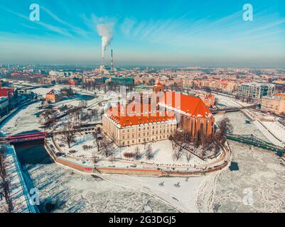 Breslau, Polen 02.15.2021 - Luftaufnahme der verschneiten Industrielandschaft mit historischen Gebäuden. Schornsteine aus dem Wärmekraftwerk im Hintergrund. Rauch verschmutzt den klaren blauen Himmel. Stockfoto