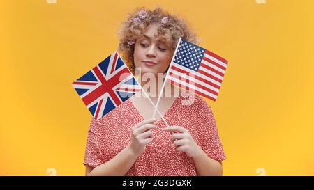 Schönes junges Mädchen, das Flaggen der USA und Großbritanniens in den Händen hält. Mädchen isoliert in gelben Hintergrund Studio. In rosa Hemd gekleidet und mit Blumen im Haar. Konzept der englischsprachigen Länder. Stockfoto