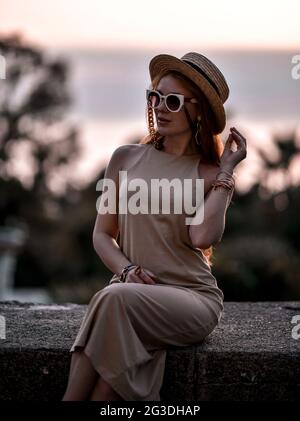Wunderschöne wohlhabende Rothaarige Frau bei Sonnenuntergang im Sommer sitzt auf Steinbrüstchen in Sonnenbrille, Strohhut und Kleid Stockfoto