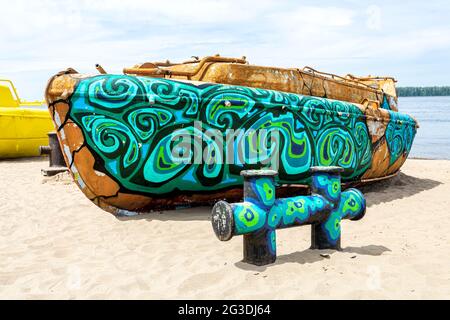 Altes rostiges Flussmotorboot mit bunten Mustern und Poller am sandigen Ufer des Flusses. Kaputtes verlassene Motorboot Stockfoto