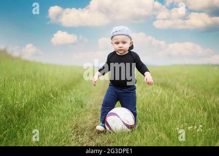 Der zweijährige kaukasische Junge mit Hut tritt auf der Wiese in den Fußball. Blauer Himmel mit weißen Wolken Hintergrund. Stockfoto