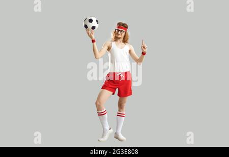 Lustiger, nerdy junger Mann mit Brille, Tanktop, Shorts und Stirnband, der den Fußball hält Stockfoto