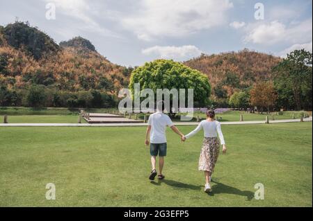 Hinter dem jungen asiatischen Paar, das im Park die Hände auf Gras und großen Baumwuchs hält Stockfoto