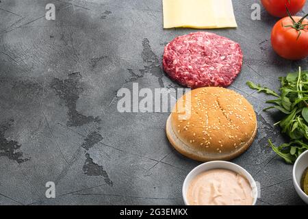 Zutaten zum Kochen von Burgern. Hacksteaks, Brötchen, Tomaten, Kräuter und Gewürze, auf grauem Steinhintergrund, mit Platz für Text Stockfoto